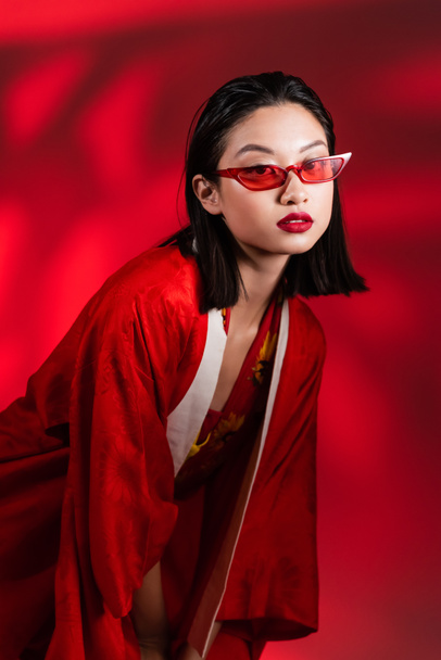 brunette asian woman in stylish sunglasses and kimono cape looking at camera on red  shaded background - Fotó, kép