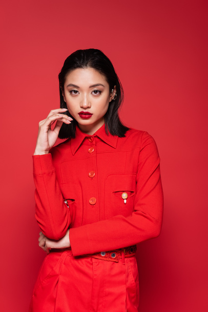 brunette asian woman in fashionable jacket holding hand near face isolated on red - Photo, Image