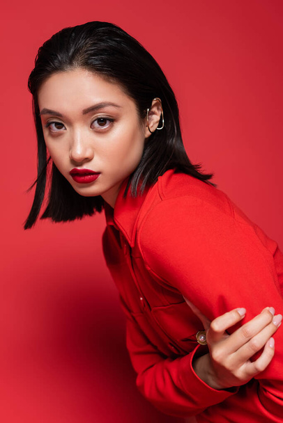 portrait of brunette asian woman with makeup and ear cuff looking at camera on red background - Fotó, kép