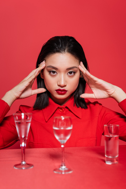 portrait of asian woman in trendy jacket posing near glasses with clear water isolated on red - Photo, Image