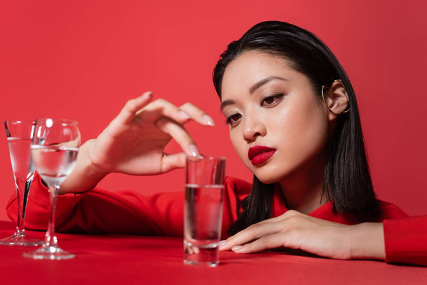 young asian woman with makeup touching glass with pure water isolated on red - Foto, afbeelding