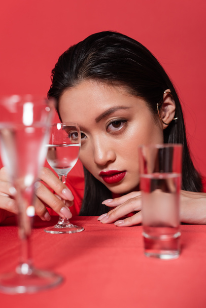 portrait of asian woman with makeup and ear cuff near glasses of clear water on blurred foreground isolated on red - Photo, Image