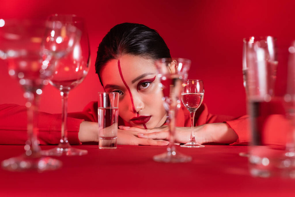 joven asiático mujer con creativo maquillaje en la cara dividido con línea cerca borrosa vasos de agua en rojo fondo - Foto, Imagen