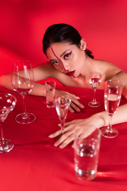 mujer asiática desnuda con maquillaje creativo posando cerca de diferentes gafas con agua sobre fondo rojo - Foto, imagen