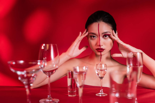 asian woman with bare shoulders and artistic makeup looking at camera near blurred glasses with water on red background - Photo, Image
