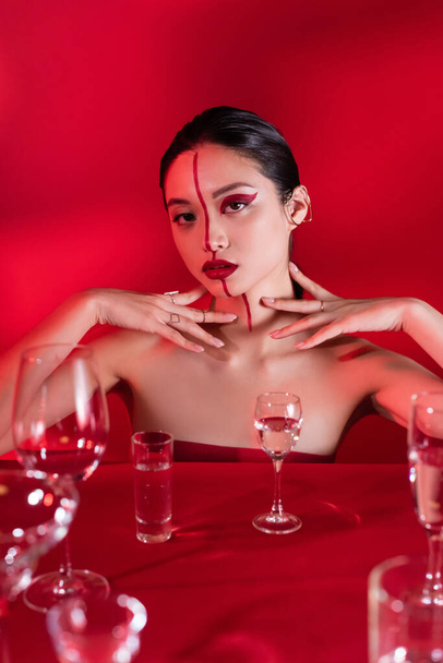stylish asian woman with bare shoulders and artistic visage touching neck near various glasses with water on red background - Foto, afbeelding