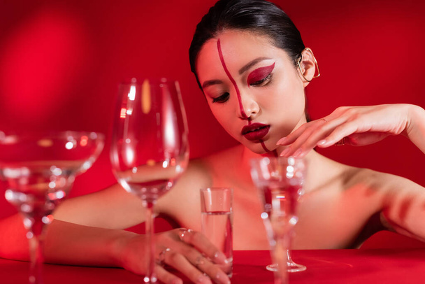 seductive asian woman with naked shoulders and artistic visage touching glass of water on red background - Photo, Image