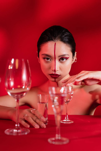 mujer asiática desnuda con maquillaje creativo y línea en la cara tocando vidrio con agua pura sobre fondo rojo - Foto, Imagen