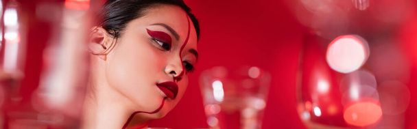 portrait of asian woman with creative makeup on face divided with line near blurred glasses on red background, banner - Fotó, kép