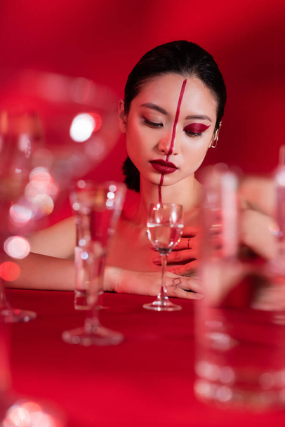 fiatal ázsiai nő piros művészi arccal nézi üveg tiszta víz elmosódott előtérben  - Fotó, kép