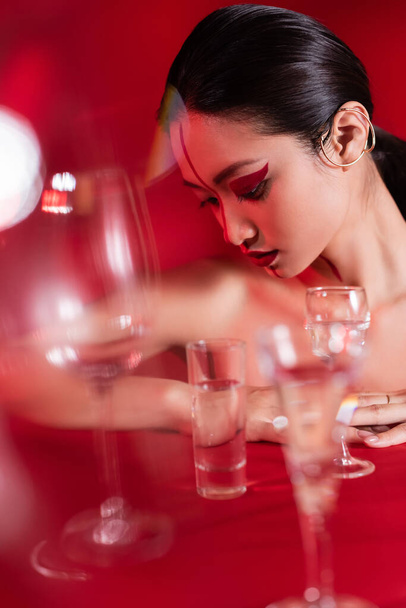 desnudo asiático mujer con oreja manguito y creativo visage cerca borrosa vasos de agua en rojo fondo - Foto, Imagen