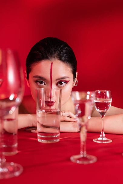 αισθησιακή Ασιάτισσα με καλλιτεχνικό μακιγιάζ κοιτάζοντας κάμερα κοντά σε γυαλιά με καθαρό νερό σε κόκκινο φόντο - Φωτογραφία, εικόνα