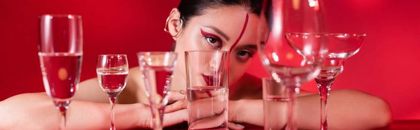 mulher asiática com visagem artística e manguito de orelha olhando para a câmera perto de óculos com água pura isolada no vermelho, banner - Foto, Imagem