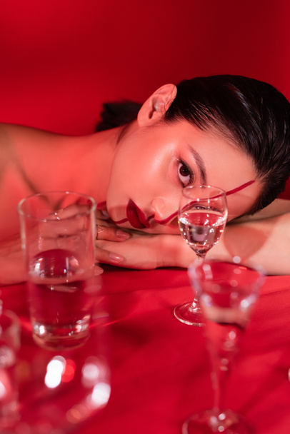 morena mujer asiática con maquillaje artístico tumbado y mirando a la cámara cerca de gafas borrosas sobre fondo rojo - Foto, imagen