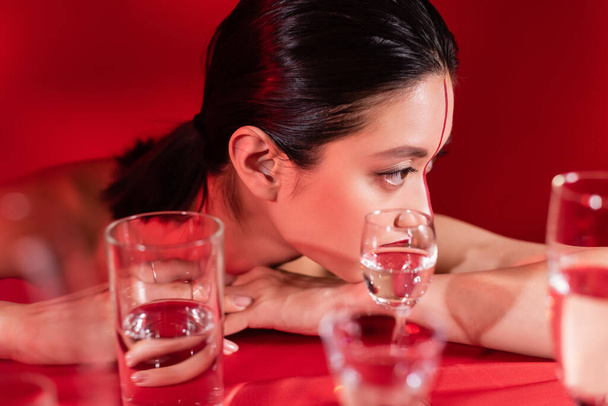 morena mujer asiática con maquillaje mirando lejos cerca borrosa vasos de agua pura sobre fondo rojo - Foto, Imagen