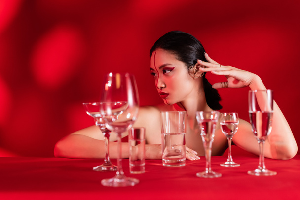 nude asian woman with creative makeup looking away near glasses with clear water on red background with shadow - Photo, Image