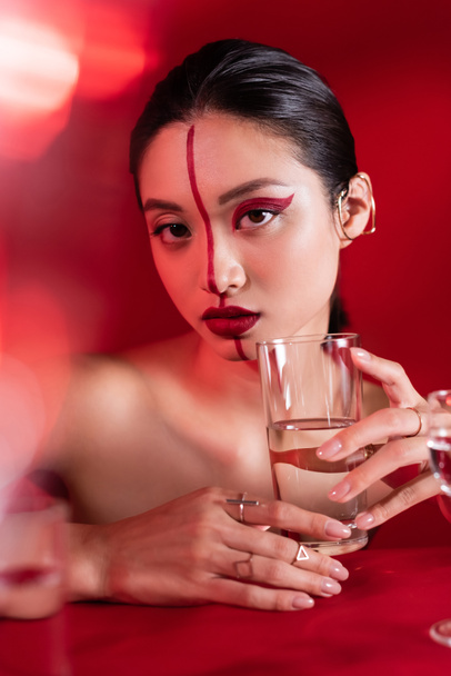 nude asian woman with red creative makeup holding glass of water and looking at camera on blurred foreground - Photo, Image