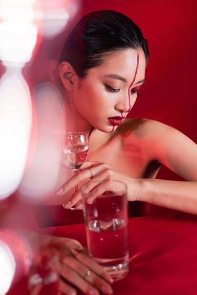 αισθησιακή Ασιάτισσα με δημιουργικό μακιγιάζ που κρατάει το ποτήρι με το νερό κοντά στην κόκκινη επιφάνεια σε θολή πρόσοψη - Φωτογραφία, εικόνα