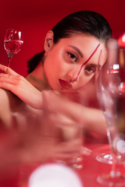 portrait de femme asiatique sensuelle avec visage artistique regardant la caméra près de lunettes floues sur fond rouge - Photo, image