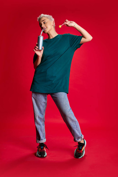 Portrait of young stylish woman with short hair posing in jeans and T-shirt, eating candy isolated over red background. Concept of youth, beauty, fashion, lifestyle, emotions, facial expression. Ad - Photo, image
