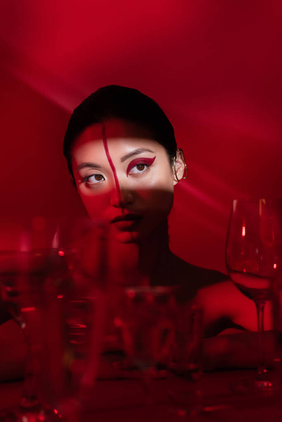 young asian woman with artistic makeup looking away in light near blurred glasses on dark red background - Photo, Image