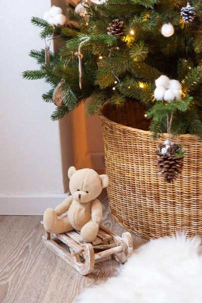 Kleiner Spielzeugbär auf einem Schlitten unter dem Ast eines Weihnachtsbaums in einem Korb. Natürliche Farben - Foto, Bild