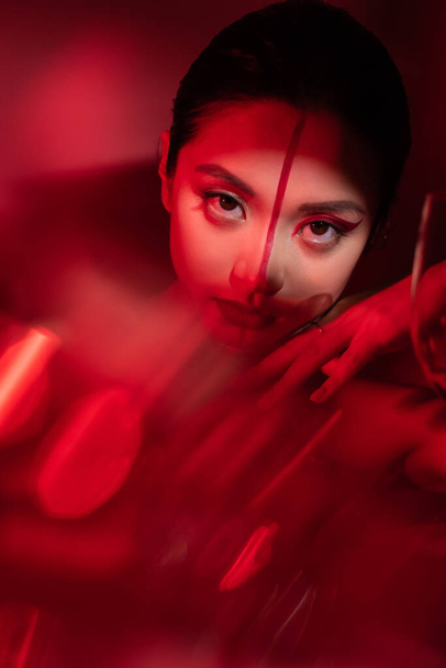 portrait of asian woman with artistic makeup looking at camera on red blurred foreground - Photo, Image
