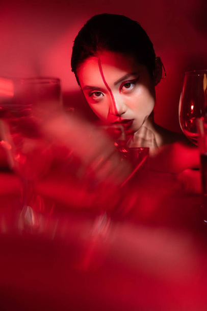 portrait of brunette asian woman with creative visage near blurred glasses on red background - Foto, afbeelding