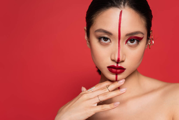 retrato de mujer asiática con rostro creativo tocando la barbilla y mirando a la cámara aislada en rojo - Foto, imagen