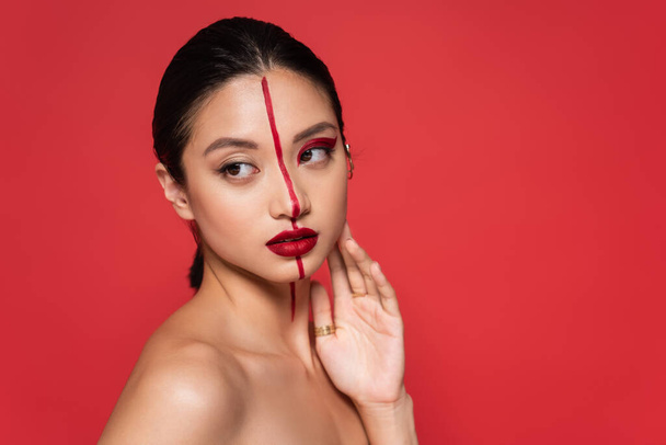 pretty asian woman with naked shoulder and artistic visage touching face while looking away isolated on red - Photo, Image