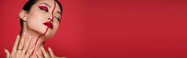 αισθησιακή Ασιάτισσα με ωτοασπίδες και δημιουργική οπτική επαφή στο λαιμό ενώ ποζάρει με κλειστά μάτια απομονωμένη σε κόκκινο λάβαρο - Φωτογραφία, εικόνα