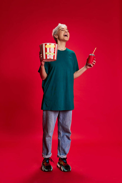 Portrait of young stylish woman with short hair posing with popcorn basket and laughing isolated over red background. Comedy. Concept of youth, beauty, fashion, lifestyle, emotions, facial expression - Foto, Bild
