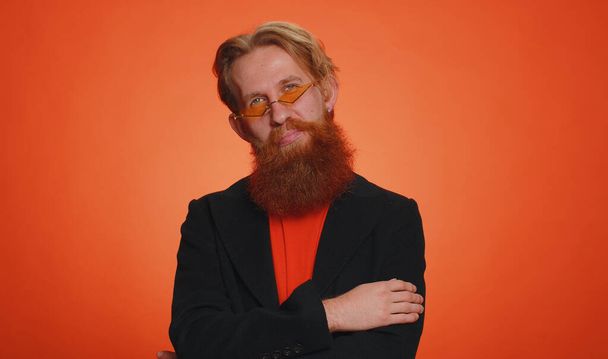 Cheerful lovely bearded ginger man in casual black jacket smiling, looking at camera. Young fashion stylish model redhead guy boy indoors studio isolated on orange wall background. Male nature beauty - Foto, immagini