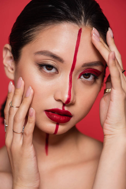portrait of brunette asian woman looking at camera while touching face with creative visage isolated on red - Foto, immagini