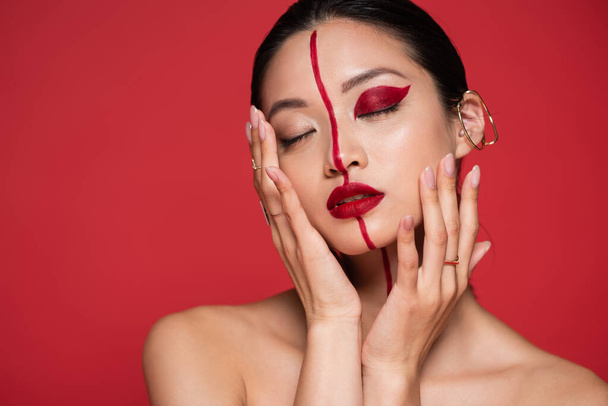 σαγηνευτική Ασιάτισσα με γυμνούς ώμους και καλλιτεχνική όψη που αγγίζει το πρόσωπο και ποζάρει με κλειστά μάτια απομονωμένη στο κόκκινο - Φωτογραφία, εικόνα