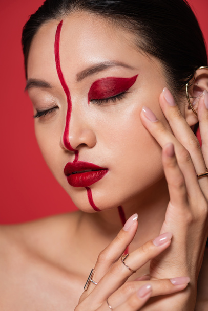 primer plano retrato de asiático mujer con cerrado ojos y creativo maquillaje tocando cara aislado en rojo - Foto, imagen