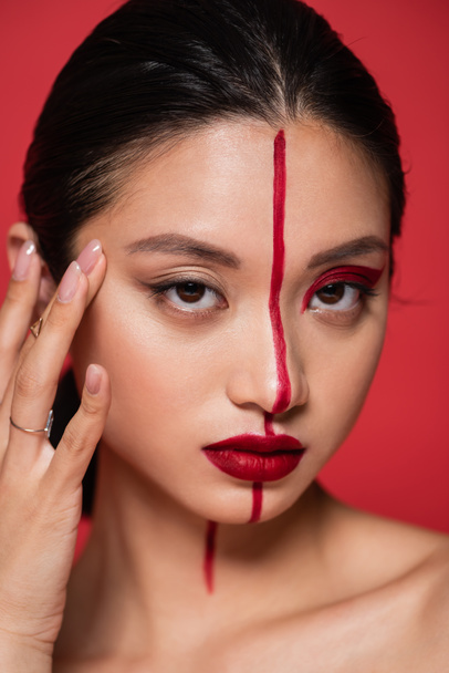 portrait of asian woman with artistic makeup touching face and looking at camera isolated on red - Foto, imagen