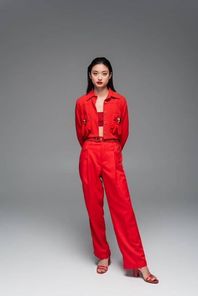 volledige lengte van trendy aziatische vrouw in rode jas en broek poseren met de handen achter rug op grijze achtergrond - Foto, afbeelding