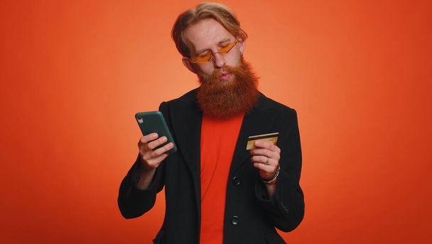 Sincere redhead man customer using credit bank card and smartphone while transferring money, purchases online shopping, payment. Finance and internet. Young guy isolated alone on orange background - Photo, Image