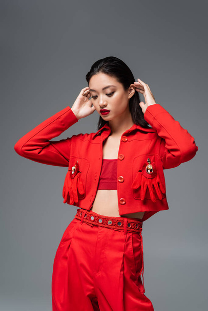 brunette asian model in red jacket decorated with brooches and gloves fixing hair isolated on grey - Zdjęcie, obraz
