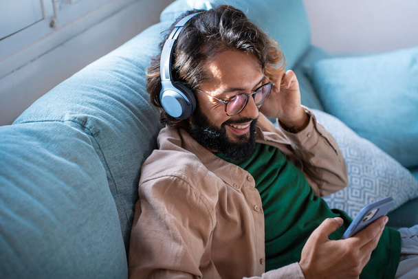 Sonriente joven con auriculares escuchar música móvil jugando en la aplicación del teléfono inteligente, chico feliz relajarse sosteniendo el teléfono disfrutar de canciones favoritas relajarse sentarse en el sofá en casa. Foto de alta calidad - Foto, Imagen