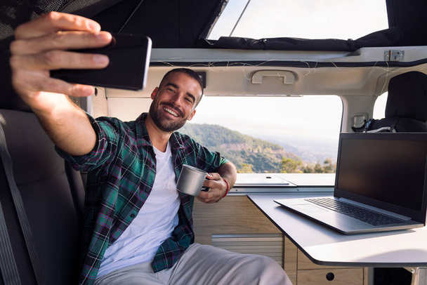 smiling young man taking a selfie inside his camper van using his mobile phone, concept of freedom and digital nomad lifestyle - Photo, Image