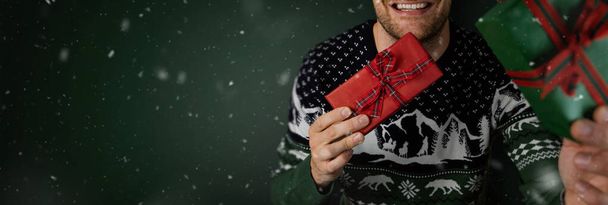 χαμογελαστός άντρας με χριστουγεννιάτικο πουλόβερ που δείχνει τυλιγμένα κουτιά δώρων σε πράσινο φόντο. banner με χώρο αντιγραφής - Φωτογραφία, εικόνα