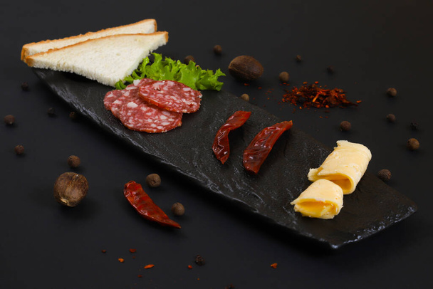Sliced smoked salami sausage with bread, vegetables and other ingredients on a black stand. Shallow depth of field - Foto, Imagem