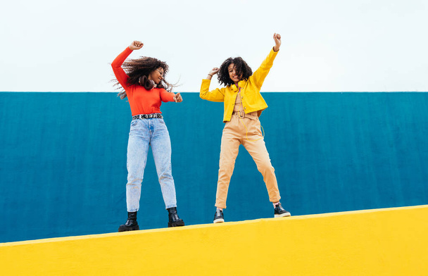 Young happy women dancing and having fun outdoor. Teenagers listening to music with smartphone and headphones in a yellow and blue modern urban area - Foto, Imagem