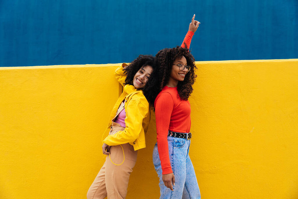 Young happy women dancing and having fun outdoor. Teenagers listening to music with smartphone and headphones in a yellow and blue modern urban area - Fotó, kép