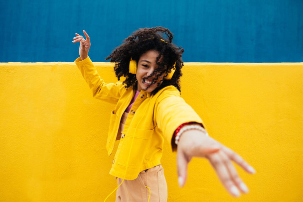 Young happy woman dancing and having fun outdoor. Teenager listening to music with smartphone and headphones in a yellow and blue modern urban area - Foto, Imagem