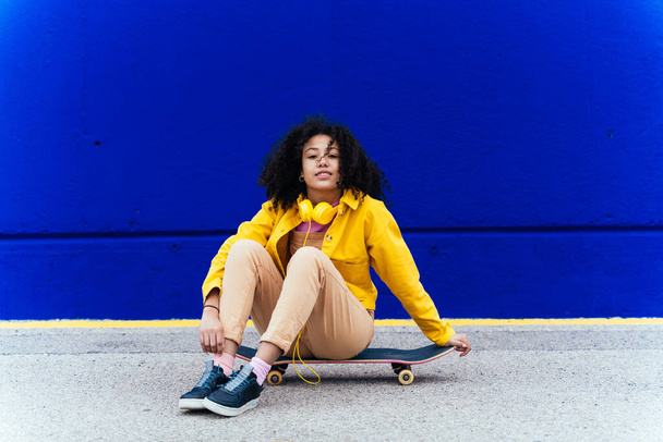 Young happy woman having fun outdoor with her skateboard. Teenager listening to music with smartphone and headphones in a yellow and blue modern urban area - 写真・画像