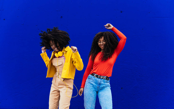 Young happy women dancing and having fun outdoor. Teenagers listening to music with smartphone and headphones in a yellow and blue modern urban area - 写真・画像
