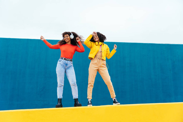 Young happy women dancing and having fun outdoor. Teenagers listening to music with smartphone and headphones in a yellow and blue modern urban area - Foto, Imagen